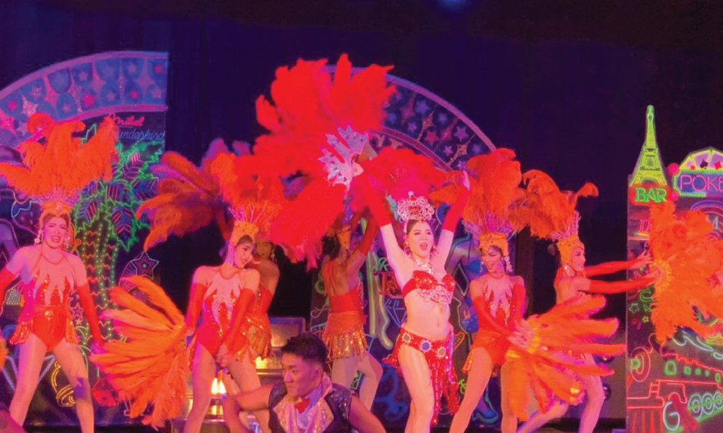 赤とオレンジのサンバ衣装を着たたくさんの女性が蛍光色を上手に使用した舞台セットの前で元気に踊っている